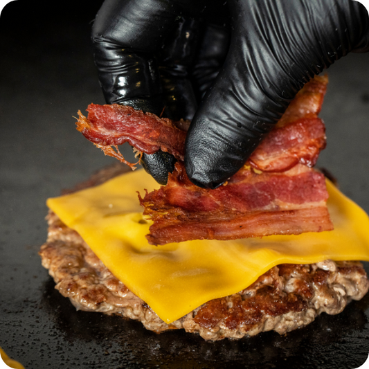 Eine behandschuhte Hand legt Streifen von gekochtem Speck auf eine Scheibe geschmolzenen Cheddar-Käse auf einem gekochten Rindfleisch-Patty auf einer schwarzen Fläche.