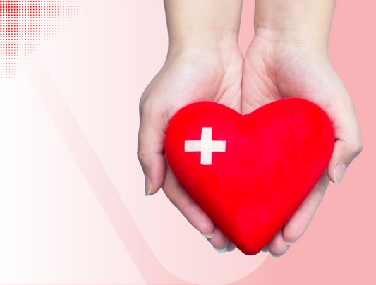Ein Paar Hände hält ein rotes herzförmiges Objekt mit einem weißen medizinischen Kreuzsymbol darauf. Der Hintergrund besteht aus einem Farbverlauf in hellrosa Tönen.