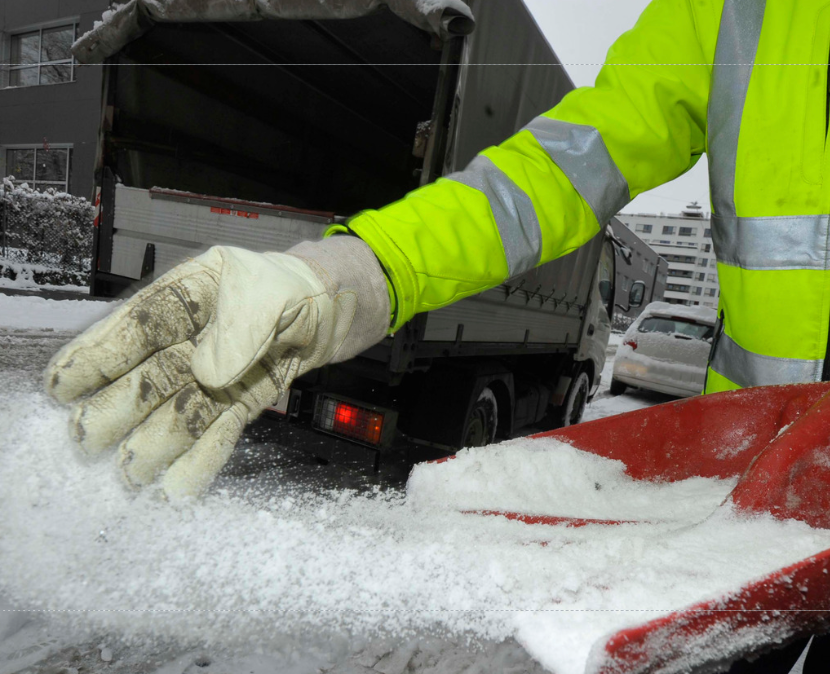 Eine Person, die eine Warnweste und Handschuhe trägt, streut mit einer roten Schaufel Salz oder Splitt auf eine verschneite Straße. Im Hintergrund sind ein Lastwagen und eine schneebedeckte Straße zu sehen.