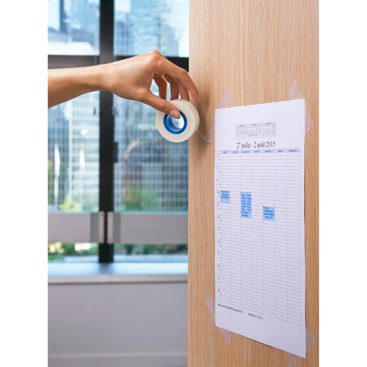 Eine Hand benutzt ein Scotch® unsichtbares Klebeband (19 mm x 33 m) von 3M Deutschland GmbH, um einen ausgedruckten Plan an einer hellen Holz-Wand zu befestigen. Im Hintergrund zeigt das Fenster den Blick auf eine urbane Stadtkulisse.