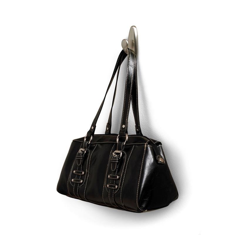 Numa bolsa de couro preta com alças duplas e fivelas decorativas pendurada num gancho de metal Command™ "clássico" da 3M Deutschland GmbH. A bolsa tem costuras brancas e uma superfície ligeiramente brilhante.