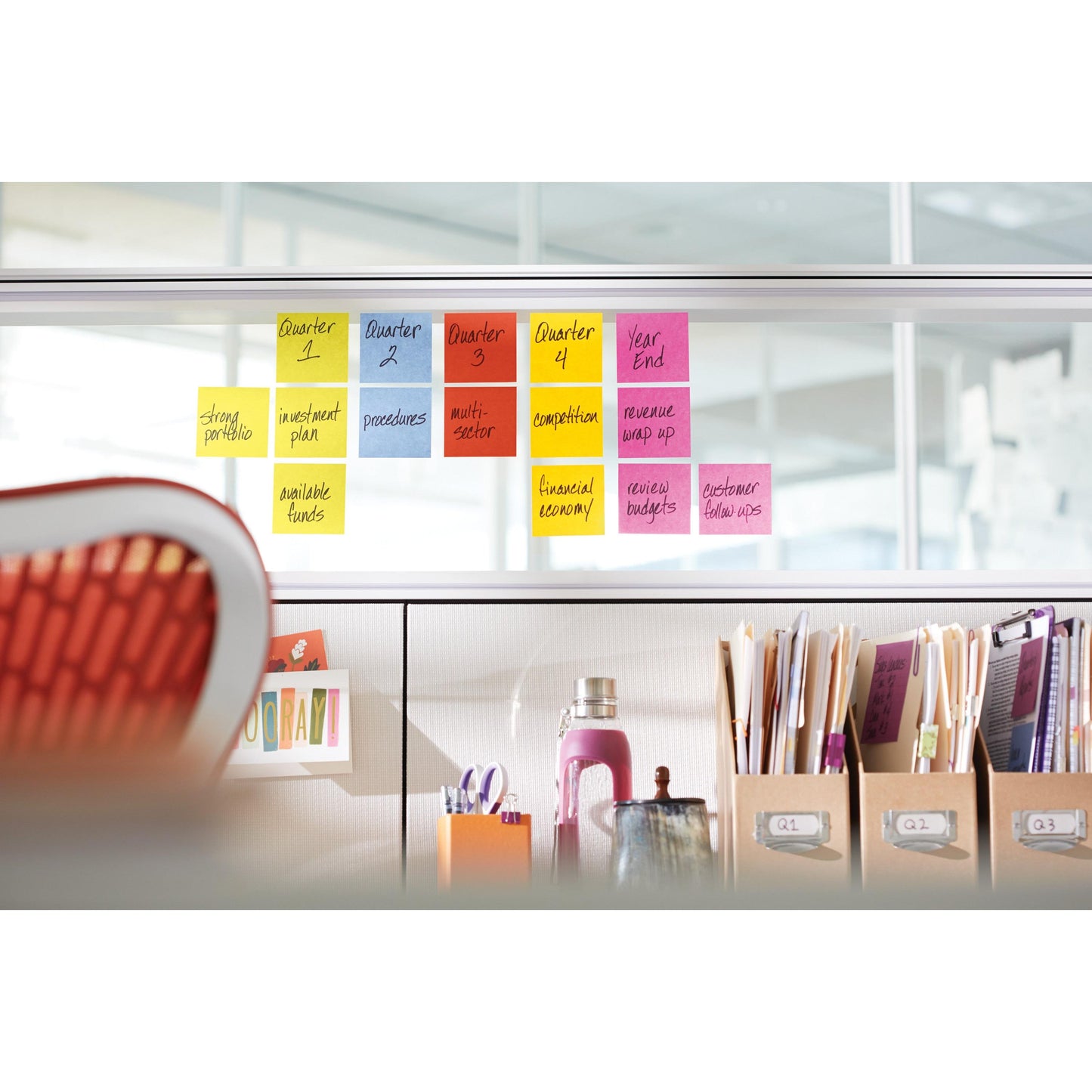 Eine Reihe farbiger Post-it® Super Sticky Würfel (3M Deutschland GmbH, 440 Blatt, 76x76 mm, 100% PEFC) zeigt auf einer Glaswand über einem Büroarbeitsplatz mit rotem Stuhl und aufgeräumten Vorräten vierteljährliche Geschäftsaufgaben an.