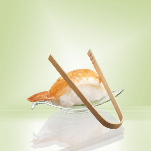 Um par de pinças para finger food de bambu de 10 cm PAPSTAR da PAPSTAR GmbH repousa sobre uma tigela de vidro ao lado de Nigiri de camarão - ideal para servir snacks ou finger food de festa - em um fundo com gradiente de cor verde pálido.