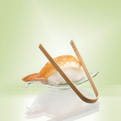Um par de pinças para finger food de bambu de 10 cm PAPSTAR da PAPSTAR GmbH repousa sobre uma tigela de vidro ao lado de Nigiri de camarão - ideal para servir snacks ou finger food de festa - em um fundo com gradiente de cor verde pálido.