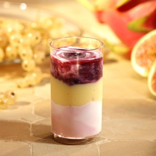 Ein Starpak 30 Fingerfood-Becher, PS 60 ml, Ø 4 cm, 7,5 cm glasklar, ist mit einem geschichtsträchtigen Dessert in Pink, Gelb und Lila befüllt und violetter Sauce garniert. Im Hintergrund: helle Beeren sowie Feigen- und Drachenfruchtscheiben.