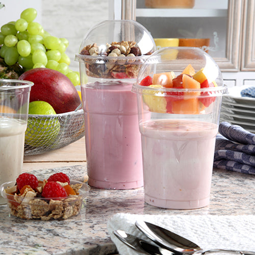 Dois parfaits de iogurte em copos de plástico, cada um fechado com uma tampa em domo Starpak 50 "Hurricane" PET redonda e transparente Ø 9,5 cm, estão sobre um balcão de cozinha – um com muesli e nozes, o outro com frutas. Ao fundo, podem-se ver bagas frescas e pratos empilhados.