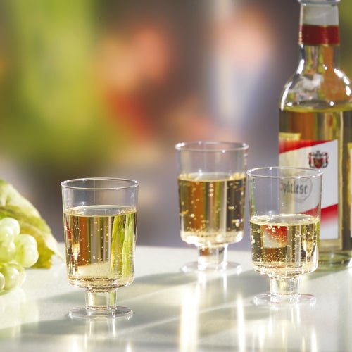 Três copos Starpak para vinho branco (PS, 0,1l, transparente) estão cheios e posicionados ao lado de uma garrafa de vinho branco e uvas verdes sobre uma superfície branca diante de um fundo desfocado.