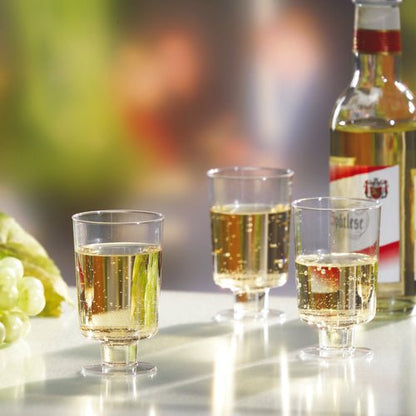 Três copos Starpak para vinho branco (PS, 0,1l, transparente) estão cheios e posicionados ao lado de uma garrafa de vinho branco e uvas verdes sobre uma superfície branca diante de um fundo desfocado.