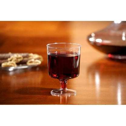 Um copo de vinho Starpak com haste para vinho tinto, PS 0,2 l transparente, está cheio sobre uma mesa de madeira; Ao fundo, snacks desfocados e uma jarra podem ser vistos.