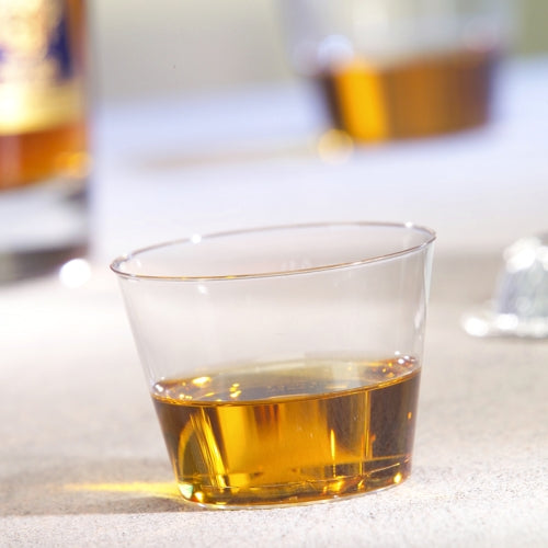 Um copo Starpak 15, PS 0,1 l cristalino, contém uma pequena quantidade de líquido âmbar sobre uma superfície clara, enquanto ao fundo um outro copo e uma garrafa estão desfocados.