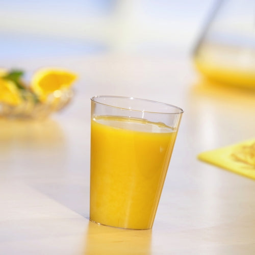 Ein Starpak 10 Trinkbecher, PS 0,2l glasklar, gefüllt mit Orangensaft steht auf einem hellen Tisch. Im Hintergrund sind unscharf Orangenscheiben und ein Krug zu sehen.
