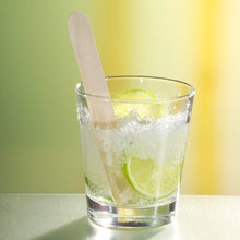 Um copo com gelo picado, fatias de lima e um PAPSTAR misturador de bebidas de madeira de 15 cm (ideal para cocktails Caipirinha) da PAPSTAR GmbH está sobre uma superfície espelhada em frente a um fundo desfocado amarelo e verde.