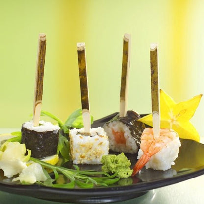 Em uma bandeja de papelão PAPSTAR (200 peças) estão três finger foods de sushi em espetos de finger food PAPSTAR (11 cm, madeira natural), acompanhados de carambola, wasabi, gengibre e verduras - perfeito para festas.