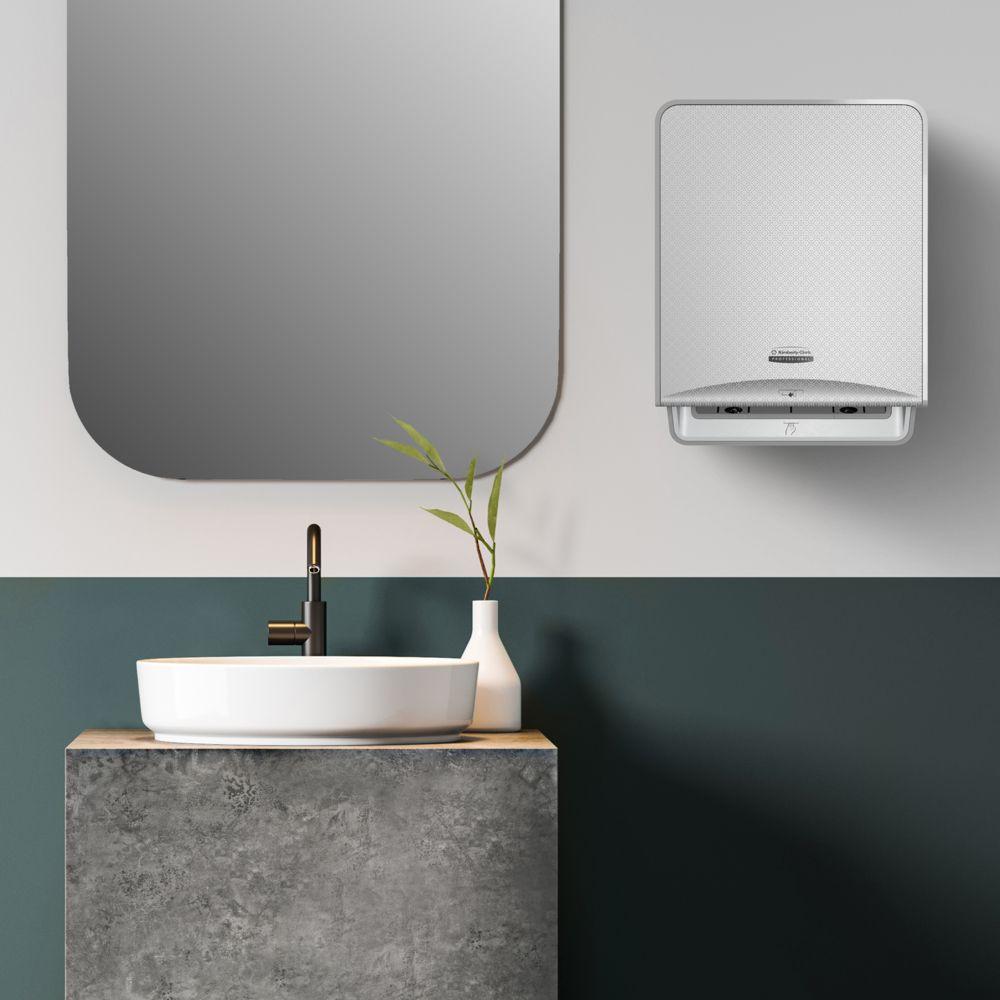 Casa de banho moderna com espelho retangular, lavatório branco sobre bancada de concreto, torneira preta, ramos verdes em um vaso e um rolo de toalha de papel Scott® Essential™ 6638 da Kimberly-Clark GmbH montado na parede.