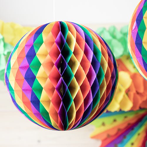 Uma aproximação da bola de favo de mel PAPSTAR 'difícil de inflamar' da PAPSTAR GmbH, uma decoração esférica em cores do arco-íris, com outra decoração colorida suavemente desfocada ao fundo.