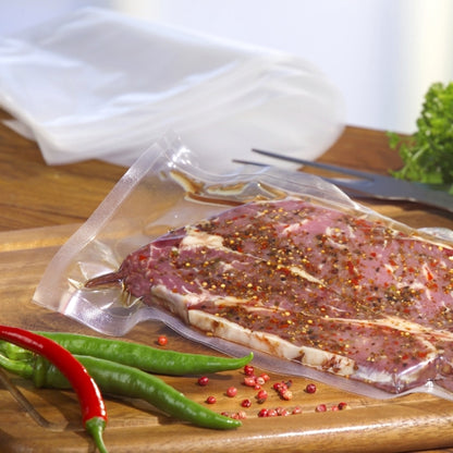 Um Starpak 100 Siegelrandbeutel, PA/PE transparente, cheio de carne marinada está sobre uma tábua de cortar de madeira entre pimentas e grãos de pimenta rosa. Verduras e utensílios completam a cena - ideal para uso com uma máquina de vácuo de câmara.
