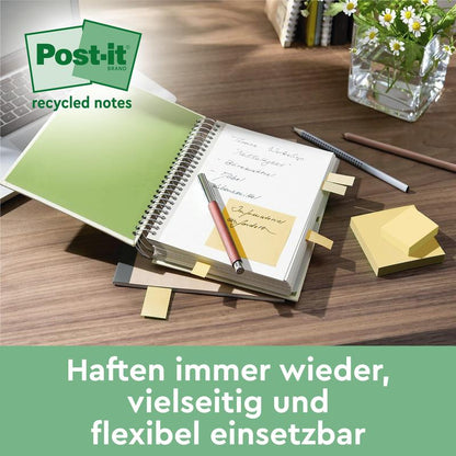 Uma mesa com um caderno aberto, vários Post-it® Super Sticky 100% Notas de Reciclagem amarelos (47,6 x 47,6 mm) da 3M Deutschland GmbH, fixados nas páginas, uma caneta, uma pilha dessas notas certificadas PEFC (70 folhas/bloco) e uma planta em vaso.