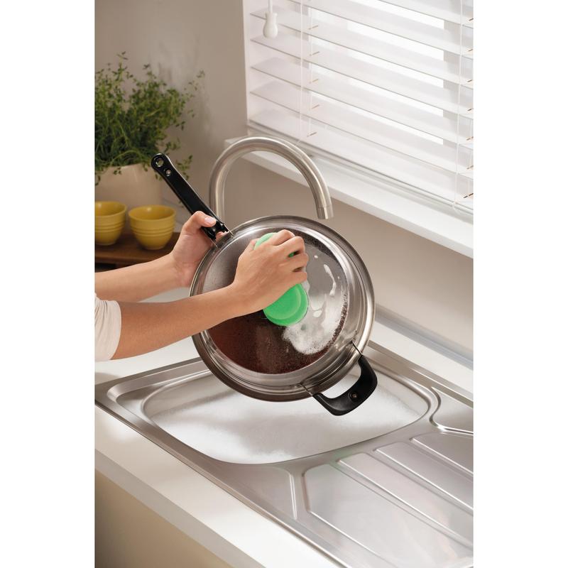 Alguém está limpando uma tampa de panela de metal sobre uma pia ensaboada com a esponja de limpeza Scotch-Brite® Classic da 3M Deutschland GmbH, perto de uma janela com persianas brancas e um balcão com uma planta e tigelas amarelas empilhadas.