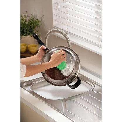 Alguém está limpando uma tampa de panela de metal sobre uma pia ensaboada com a esponja de limpeza Scotch-Brite® Classic da 3M Deutschland GmbH, perto de uma janela com persianas brancas e um balcão com uma planta e tigelas amarelas empilhadas.