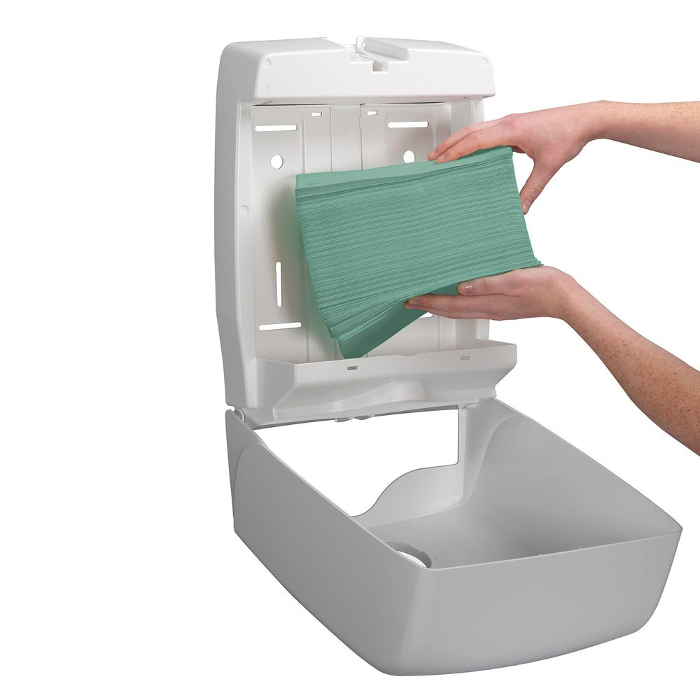 Uma pessoa coloca uma pilha de toalhas Hostess™ NATURA™ da Kimberly-Clark GmbH - dobragem em ziguezague, Pequena (23,00 cm x 25,00 cm) num dispensador de toalhas de papel branco montado na parede.