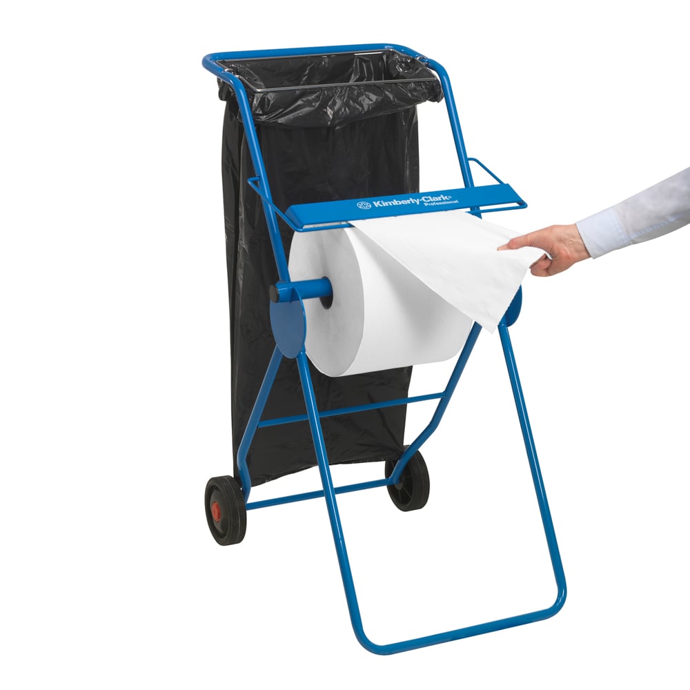 Um dispensador de toalhas de papel azul com um grande rolo de papel médico branco Scott® ESSENTIAL™ (6 x 200 folhas) da Kimberly-Clark GmbH destaca a higiene; um saco de lixo preto está próximo, enquanto a mão de uma pessoa puxa uma folha do rolo.