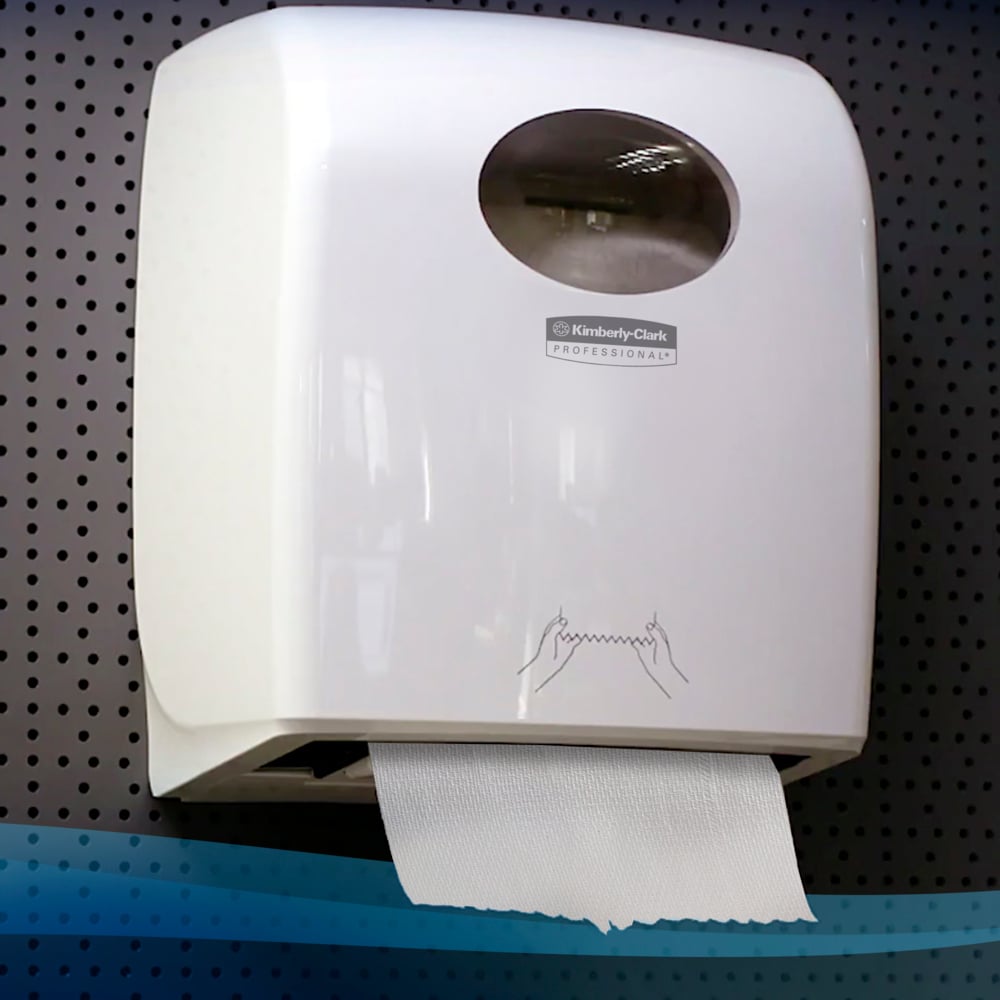 Um dispensador de parede branco com janela transparente contém um rolo de toalhas de mão Scott® (304,00 m x 19,80 cm) da Kimberly-Clark GmbH com uma folha pendurada sobre uma parede cinza perfurada.