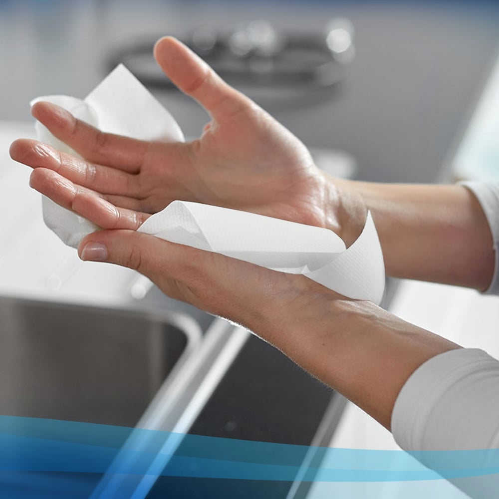 Uma pessoa limpa as mãos com um rolo de toalhas de mão Scott® branco (304,00 m x 19,80 cm) da Kimberly-Clark GmbH sobre uma pia, com a bancada e a pia visíveis ao fundo.