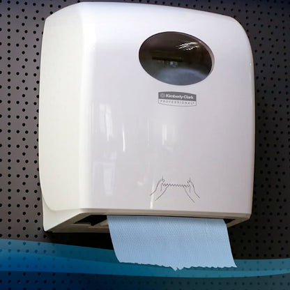Um dispensador branco montado na parede da Kimberly-Clark GmbH contém um rolo de toalhas de mão Scott® (304,00 m x 19,80 cm) com uma toalha azul e um guia ilustrado simples diante de um fundo escuro de chapa perfurada.
