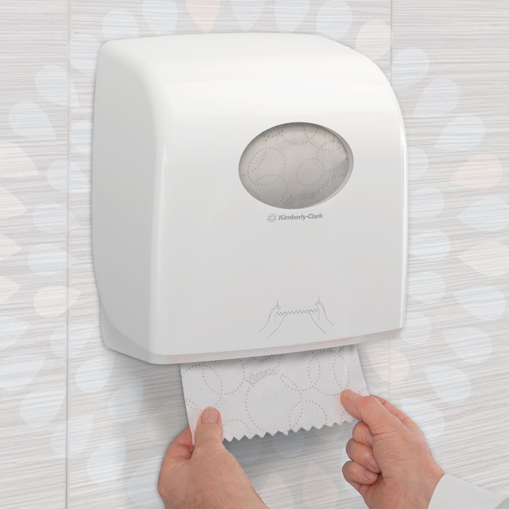 Alguém puxa uma toalha de mão Kleenex® Ultra™ Slimroll™ de um dispensador branco montado na parede com janela de visualização e símbolos em um banheiro azulejado. Produto: caixa de 6 rolos da Kimberly-Clark GmbH.