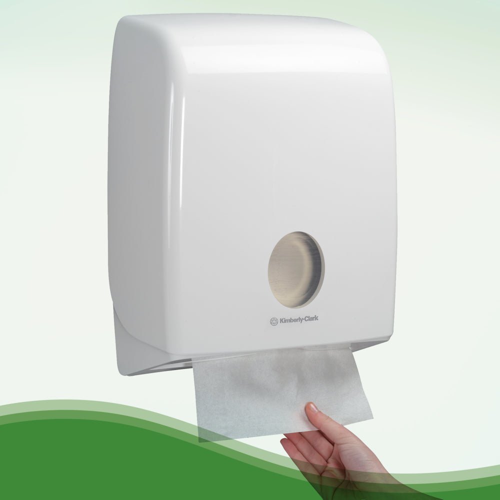 Uma mão puxa toalhas Hostess™ NATURA™ - dobragem em ziguezague, Pequena (23 x 25 cm) da Kimberly-Clark GmbH de um dispensador de parede branco com janela redonda na frente.