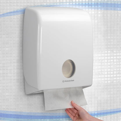 Uma mão puxa uma toalha de papel de um dispensador Aquarius™ branco montado na parede para toalhas de mão dobradas com Dobra em C da Kimberly-Clark GmbH, instalado em uma parede azulejada.