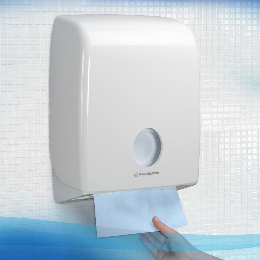 Uma mão puxa uma toalha dobrada azul Scott® Control™ Interfold de um dispensador branco da Kimberly-Clark GmbH, com a caixa contendo 15 pacotes de 240 folhas cada, sobre um fundo com azulejos.