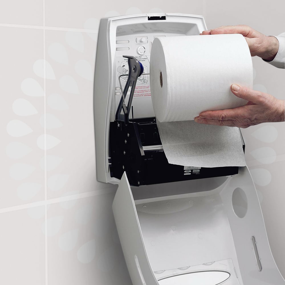 Eine Person lädt eine Kleenex® Ultra™ Handtücher - Rolle der Kimberly-Clark GmbH in einen weißen, offenen Wandspender in einer Toilette ein, um ein hygienisches Händetrocknen zu gewährleisten. Im Deckel des Spenders ist eine Anleitung aufgedruckt.