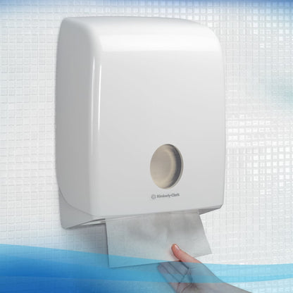 Uma mão puxa uma toalha de mão Scott® Control™ Interfold de um dispensador branco da Kimberly-Clark em um lavatório com azulejos, garantindo a secagem higiênica das mãos.