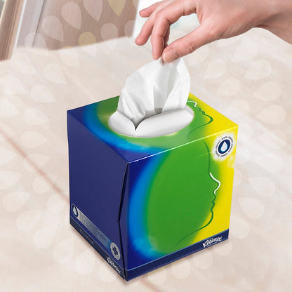 Uma mão puxa um lenço da caixa de cubos de lenços cosméticos Kleenex® / Branco da Kimberly-Clark GmbH com design em azul, verde e amarelo e uma abertura oval branca. Um perfil estilizado sugere cuidado facial diante de um fundo suavemente desfocado.