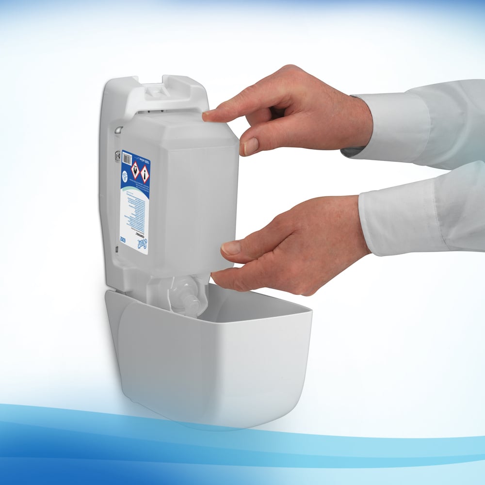 Uma pessoa está a encher um dispensador montado na parede com um cartucho de 1 litro de Espuma Desinfetante para as Mãos à Base de Álcool Scott® CONTROL da Kimberly-Clark GmbH e exibe o sinal de aviso. As suas mãos são visíveis e ela está a usar uma camisa branca de mangas compridas.