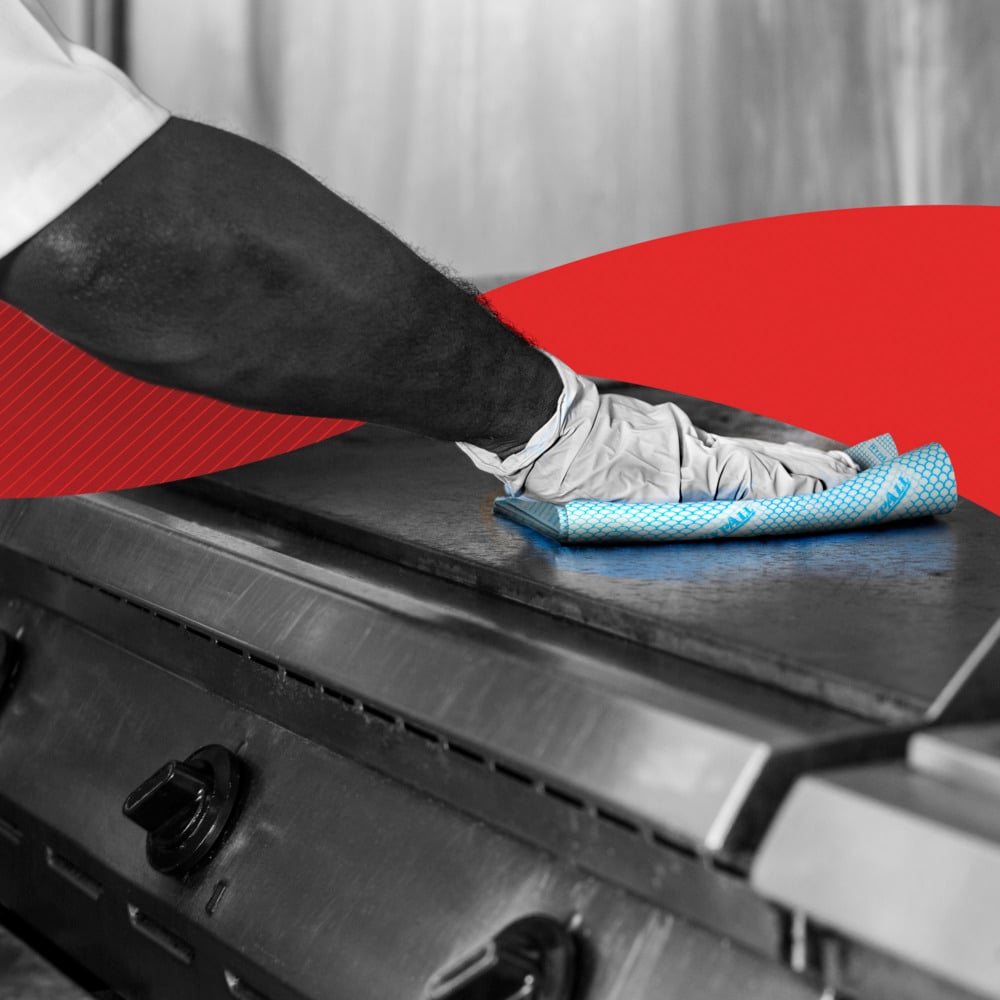 Uma pessoa com luvas limpa uma placa de fogão de metal com um pano de limpeza azul WypAll® X50 - Interfold (41,80 cm x 24,70 cm) da Kimberly-Clark GmbH. Um gráfico vermelho curvo no fundo destaca a limpeza e o cuidado da cozinha.