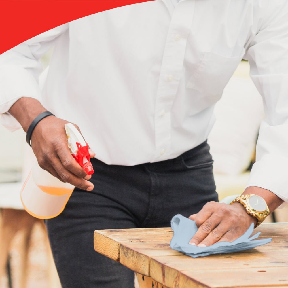 Uma pessoa com camisa branca e relógio limpa uma mesa de madeira com um pano azul, possivelmente WypAll® L20 General Clean - caixa BRAG™ da Kimberly-Clark GmbH. A imagem tem uma borda vermelha curva na parte superior, destacando a atenção aos detalhes.