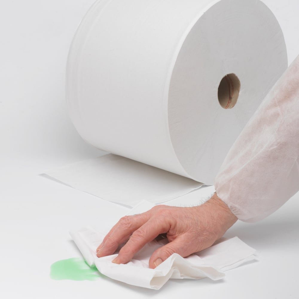 Eine Hand wischt einen grünen Fleck mit saugfähigem Papier auf, während im Hintergrund eine WypAll® L20 General Clean Großrolle (2 Rollen x 1085 Blatt) von Kimberly-Clark GmbH auf einer weißen Fläche liegt, bereit für schwierige Reinigungsarbeiten.
