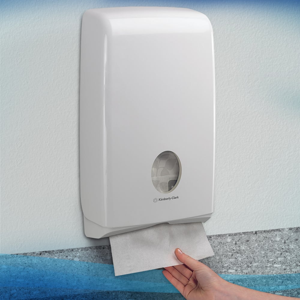 Uma mão puxa uma toalha Scott® SLIMFOLD de um dispensador de parede branco da Kimberly-Clark GmbH com uma janela de visualização, que mostra toalhas dobradas no interior. O dispensador higiênico está montado em uma parede clara; ele comporta 16 clips x 110 folhas por caixa.