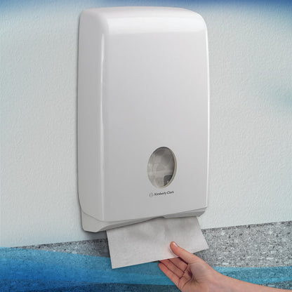Uma mão puxa uma toalha Scott® SLIMFOLD de um dispensador de parede branco da Kimberly-Clark GmbH com uma janela de visualização, que mostra toalhas dobradas no interior. O dispensador higiênico está montado em uma parede clara; ele comporta 16 clips x 110 folhas por caixa.