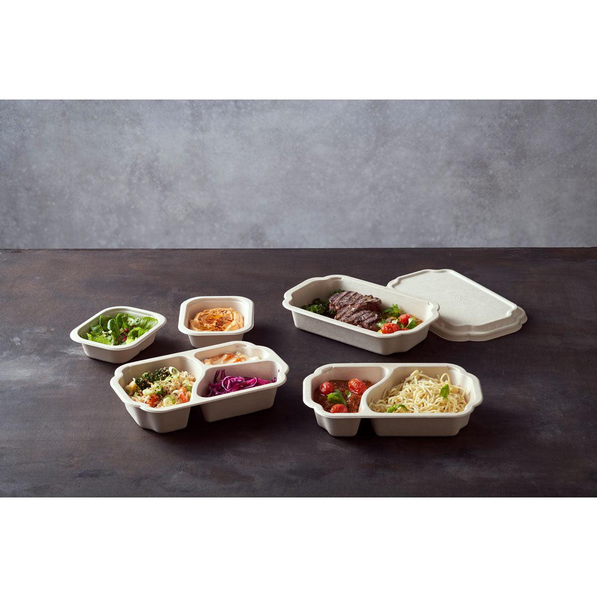 Numa superfície escura encontram-se vários recipientes de bagasse «Tray Side Dish Fibre 1-Comp 55 mm Natur Coated» da Duni GmbH, cada um dos quais contém pratos diferentes; uma embalagem com 320 unidades contém um recipiente com a tampa parcialmente aberta.