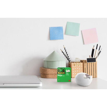 Uma secretária com um laptop fechado, um dispensador de mesa Scotch™ Karim Rashid da 3M Deutschland GmbH e uma caixa de fita adesiva, recipientes para canetas e lápis, um pequeno ornamento em forma de casa e três notas adesivas coloridas na parede branca.