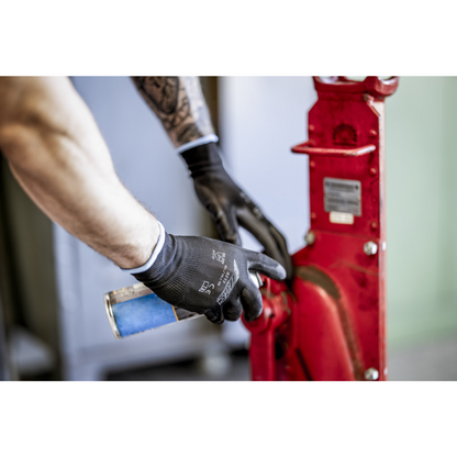 Uma pessoa com luvas de nylon NITRAS pretas, revestidas com PU da AS Arbeitsschutz GmbH, está a limpar ou lubrificar com uma lata de spray azul uma peça de máquina vermelha de metal. Apenas braços e mãos são visíveis.