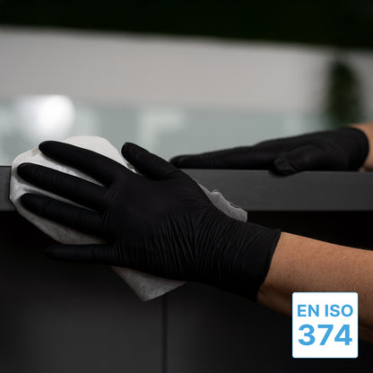 Uma pessoa que usa as luvas descartáveis extra grossas ARNOMED NITRIL BLACK XTRA STRONG de nitrilo preto da ARNOWA GmbH limpa uma superfície escura com um pano branco. No canto inferior direito aparece a etiqueta azul e branca "EN ISO 374".