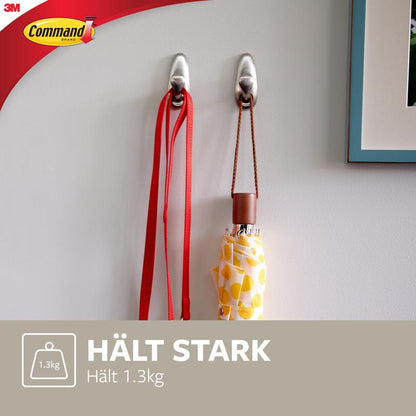 Dois ganchos de metal Command™ "clássico" da 3M Deutschland GmbH estão montados numa parede branca e seguram uma trela vermelha e um guarda-chuva amarelo com padrões. O texto diz: "SEGURA FORTEMENTE Suporta 1.3kg" e mostra o logotipo Command.