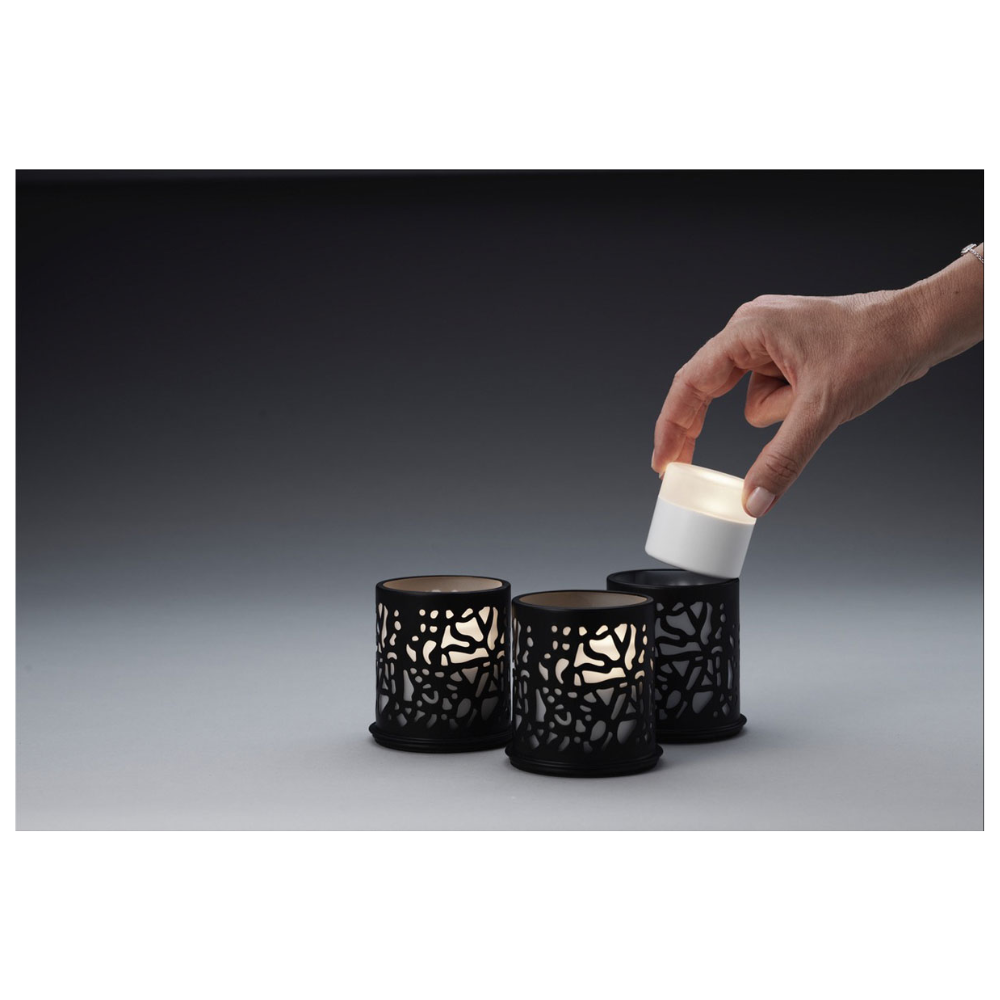 Uma mão coloca uma pequena vela cilíndrica em um dos três castiçais Twine 75 x Ø 75 mm Preto da Duni GmbH, que possuem recortes elaborados e são ideais para design moderno. A decoração de mesa está posicionada diante de um fundo com gradiente de cinza escuro para cinza claro.