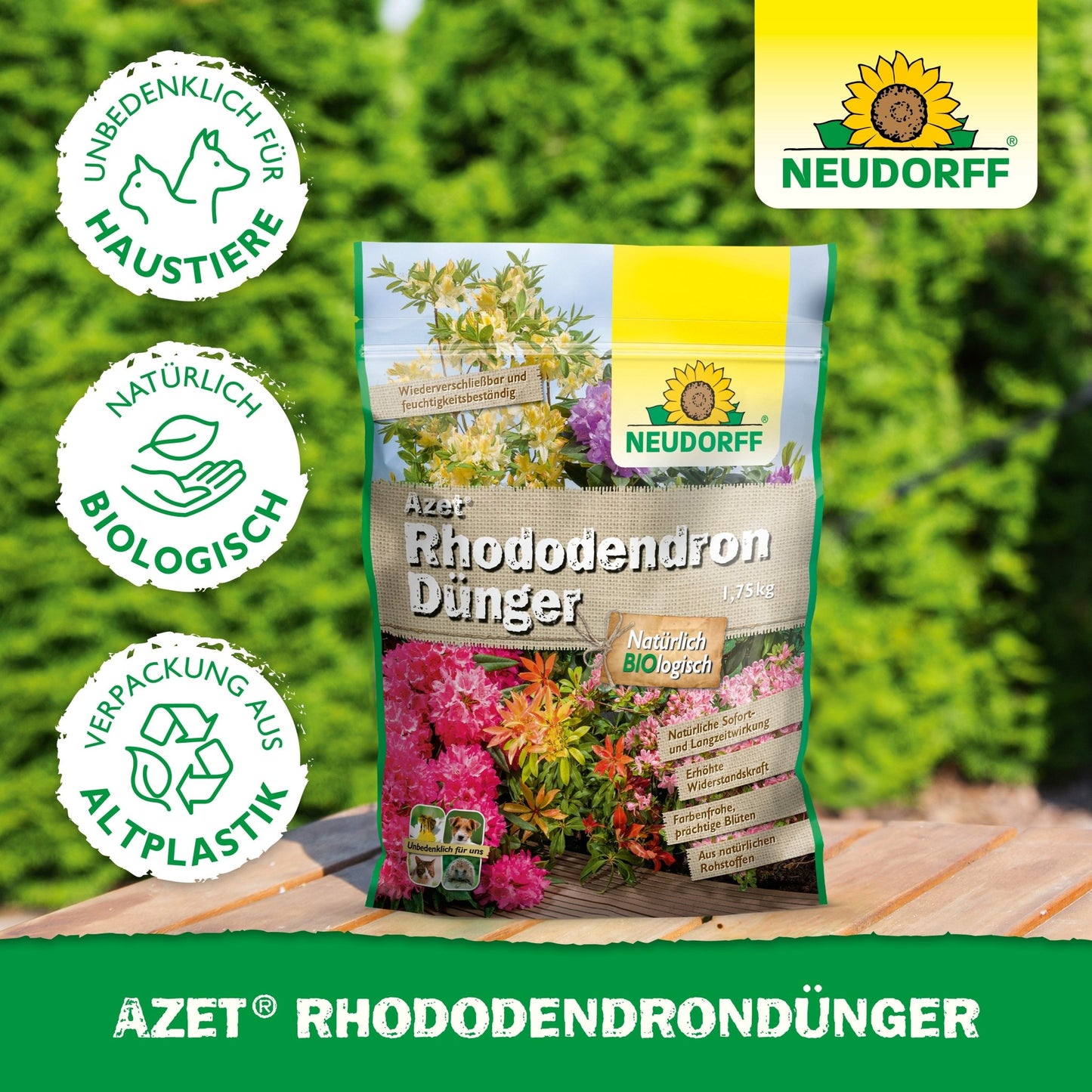Um saco de Azet RhododendronDünger da Neudorff Shop está sobre uma superfície de madeira ao ar livre em frente a sebes verdes. Na embalagem de bioplástico, está escrito que é biológico, amigo dos animais de estimação e ideal para rododendros e azáleas.