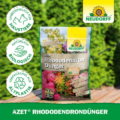 Um saco de Azet RhododendronDünger da Neudorff Shop está sobre uma superfície de madeira ao ar livre em frente a sebes verdes. Na embalagem de bioplástico, está escrito que é biológico, amigo dos animais de estimação e ideal para rododendros e azáleas.