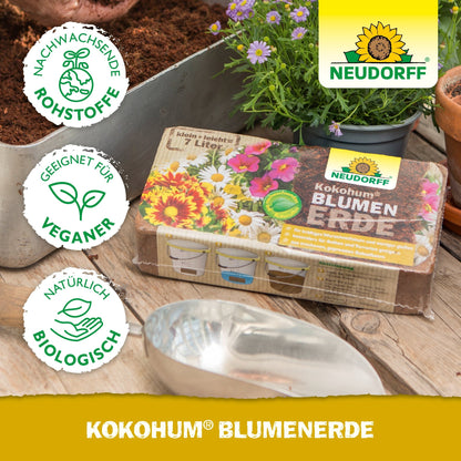 Uma embalagem de Neudorff Shop Kokohum BlumenErde está sobre uma mesa com terra, uma pá de metal, vasos de flores e plantas. Os símbolos mostram que o produto é vegano, biológico e feito de materiais renováveis.